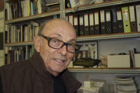 An older balding man with glasses and a brown jacket stands in front of shelves of files. Leo Christie