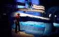 A photo of a mini golf installation featuring a large blue wave. A person is standing in front of it, putter in hand. An airplane installation with a flag “Waves of Fortune’ is hovering just above the wave.