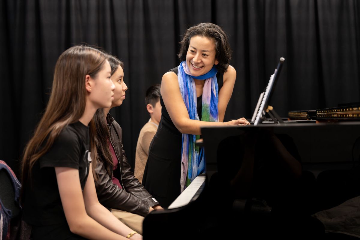 Woman leans on grand piano smiling, where two students sit and look up at her. Strike a Chord.