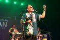 A man on stage with his arm in the air. He is wearing a green print shirt. There are drummers behind him.