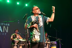 A man on stage with his arm in the air. He is wearing a green print shirt. There are drummers behind him.