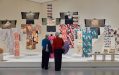 Two elderly woman looking at a display of a dozen kimonos hanging in a gallery with white walls.
