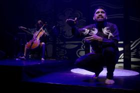 A man is crouching on stage, a cellist is behind him.
