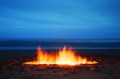 A moody and dramatic image by photographer Caterina Pacialeo of a firepit on a beach at dusk. Flames dance towards the cloudy horizon. Sydney Contemporary 2025 expands with the addition of the inaugural Photo Sydney.