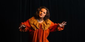 A woman with dark hair with her arms outstretched. She's wearing an elaborate burnt orange costume.
