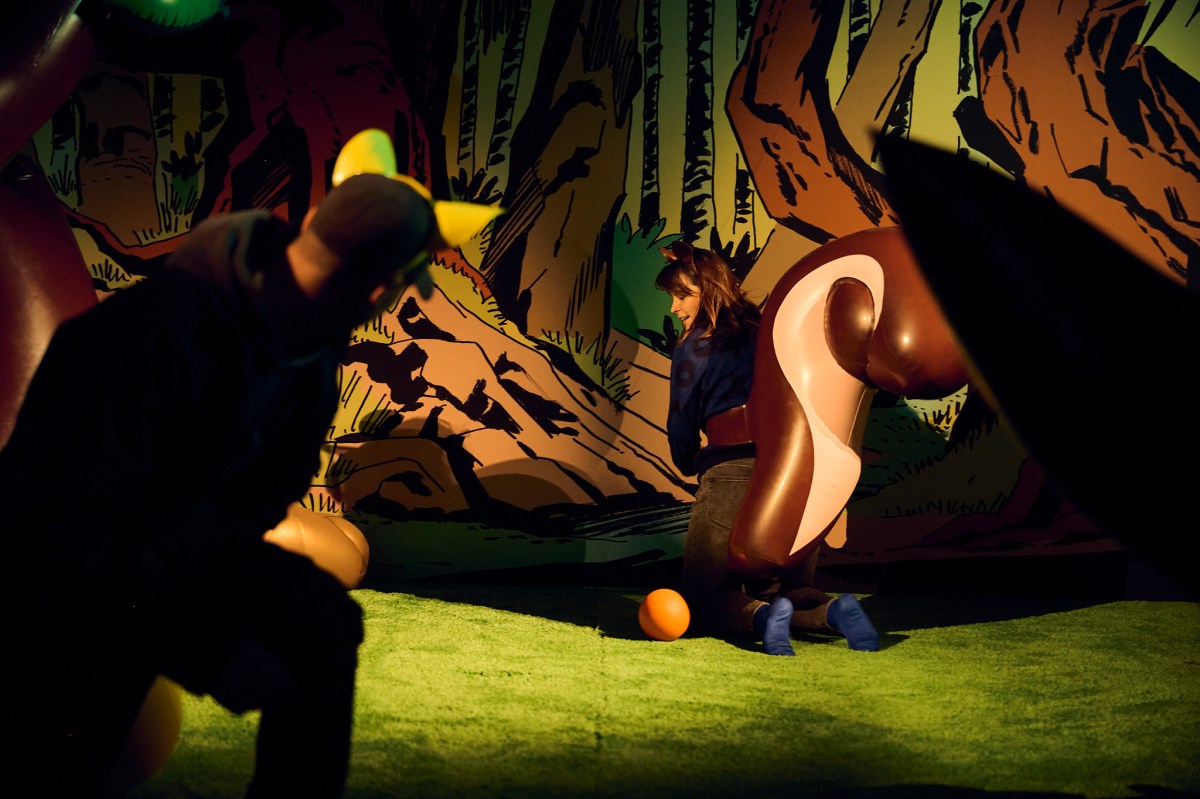 A man and a woman wearing large blow-up latex tails attempt to use them as putters in a fantasy woodland game of mini golf. Swingers
