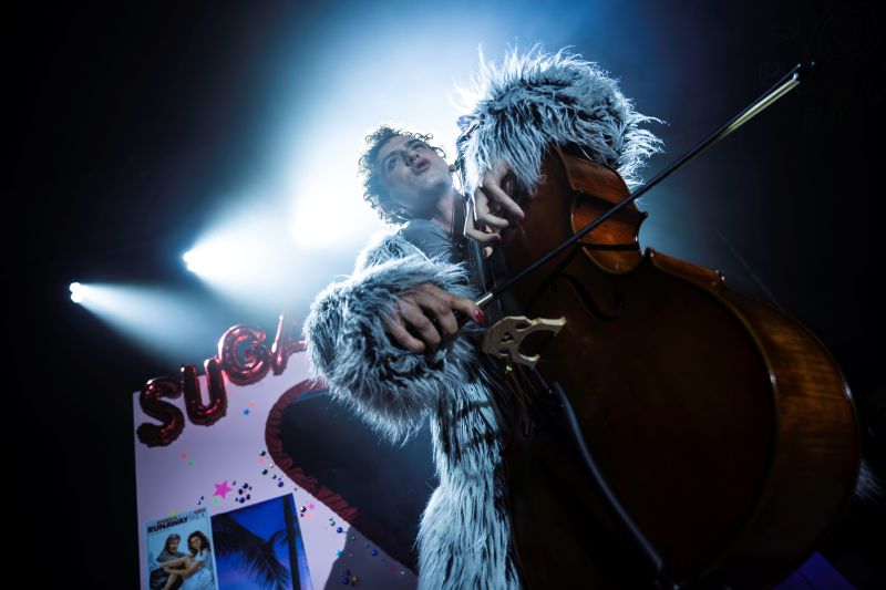 Tomas Kantor's 'Sugar' has been programmed for Adelaide Cabaret Festival 2025. The photo, taken from a low angle, looks up to show Kantor playing a cello with a bow. He is wearing a grey fau-fur coat and his lips are pursed as he sings; his curly hair is visible in the stage lights shining down from above.