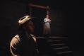 A young man in a WWI soldier's uniform sits in profile in the dark. In the background is a woman in white.