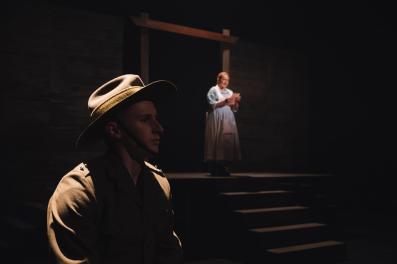 A young man in a WWI soldier's uniform sits in profile in the dark. In the background is a woman in white.