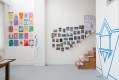 A photograph of a brightly lit artist's studio. Art adorns the white walls, including a bold geometric design in blue and multiple sketches and colourful drawings. A flight of wooden stairs leads tantalisingly upwards, out of sight.