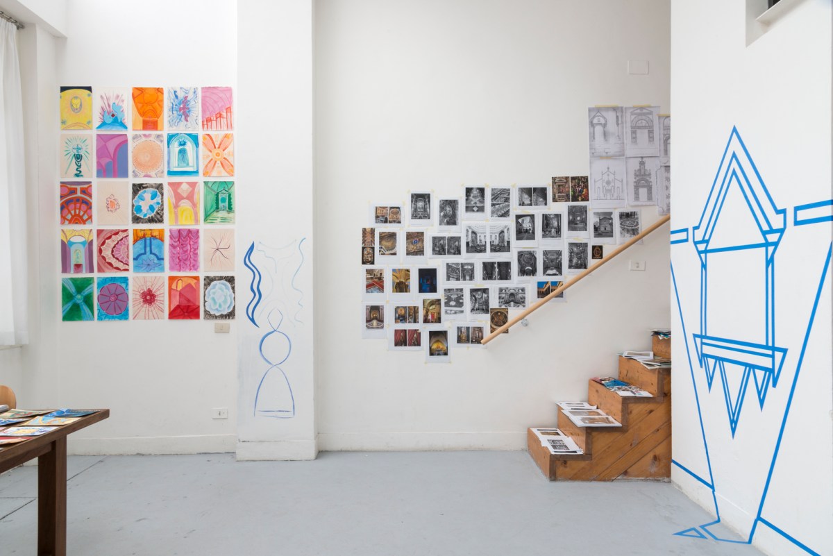 A photograph of a brightly lit artist's studio. Art adorns the white walls, including a bold geometric design in blue and multiple sketches and colourful drawings. A flight of wooden stairs leads tantalisingly upwards, out of sight.