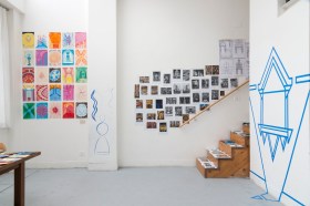 A photograph of a brightly lit artist's studio. Art adorns the white walls, including a bold geometric design in blue and multiple sketches and colourful drawings. A flight of wooden stairs leads tantalisingly upwards, out of sight.