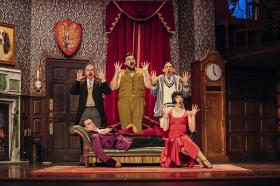 In an elaborate drawing room, three men and one woman have their arms up and mouths open in suprise. One man is lying on a chaise longue