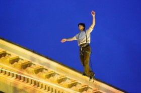 a tightrope walking performer scaling the edge of a high building ledge against a bright blue sky.
