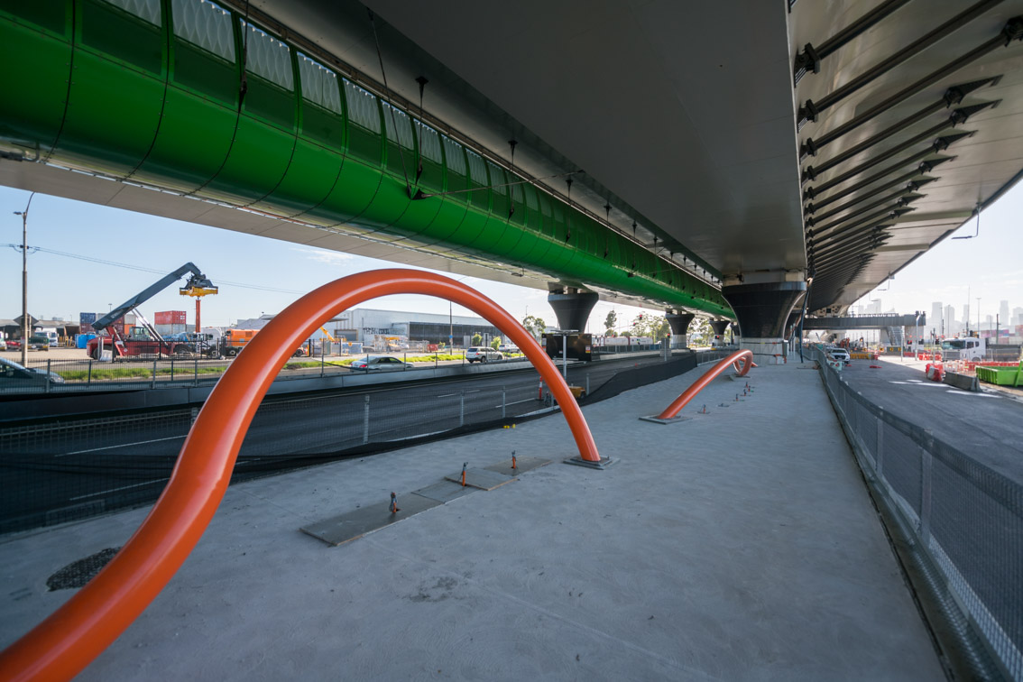An orange curvy barrier under a green raised tube under a freeway, designed to carry bicycles. Veloway, Open House Melbourne 2025