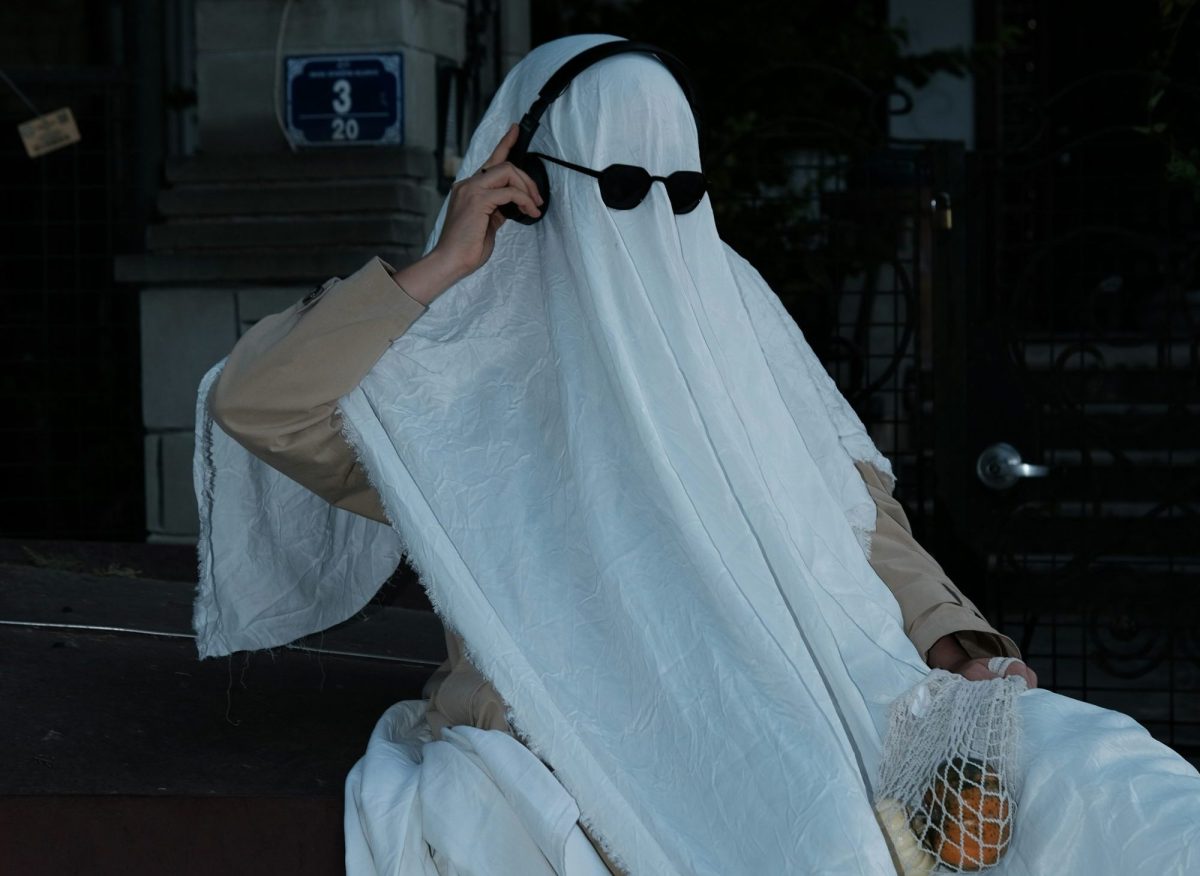 A person in a ghost costume with sunglasses and headphones is on the phone.