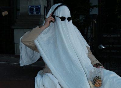 A person in a ghost costume with sunglasses and headphones is on the phone.