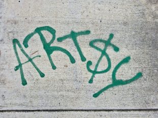 Green spraypaint reads 'ART$Y' on grey pavement - a reflection of arts funding.