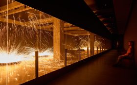An installation at Mona (Museum of Old and New Art) where molten steel drips from the ceiling and create massive sparks. Some visitors look on from a bench.