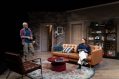 A living room scene. There is a man standing up looking at a woman seated on a brown couch.