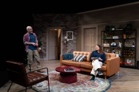 A living room scene. There is a man standing up looking at a woman seated on a brown couch.