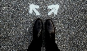 two black shoe clad feet with arrows facing in opposing directions on the ground. visual arts public sector