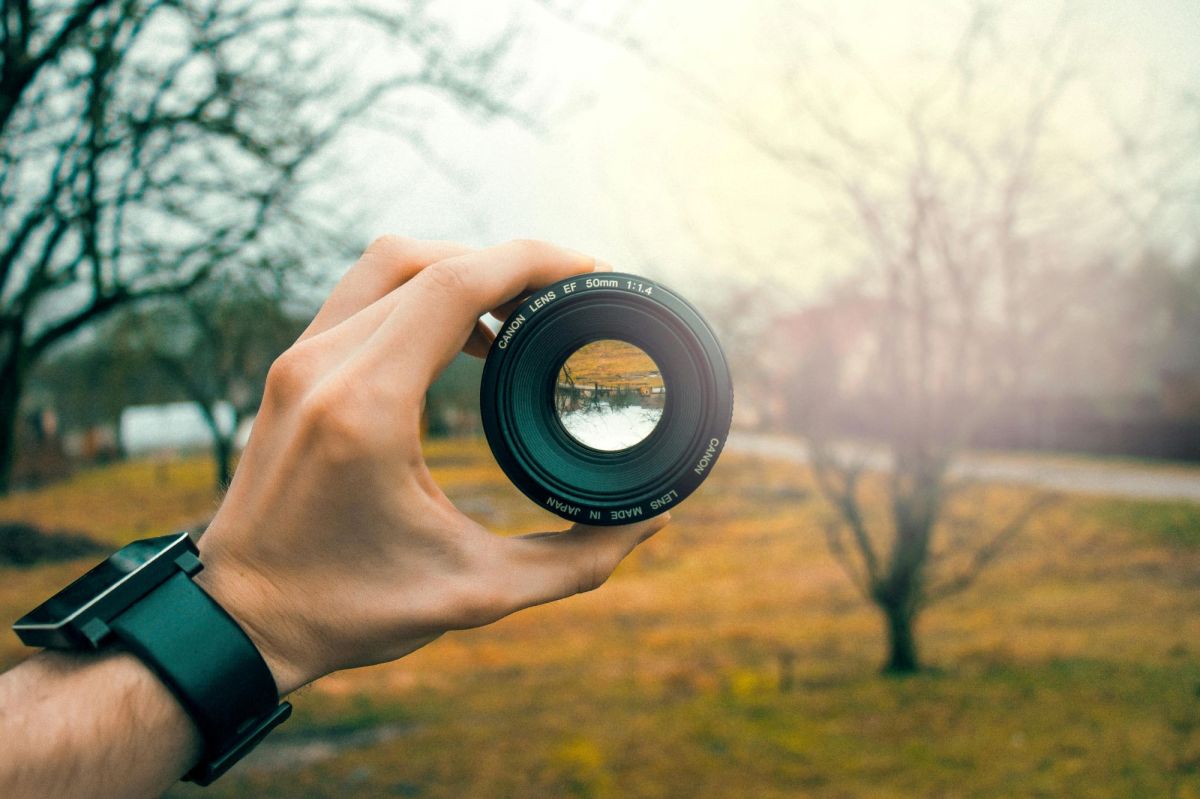 A photograph of a fair-skinned hand holding up a camera lens in front of a stark, misty winter landscape of bare trees, moorland and a river. The landscape is reversed in the lens. The photo is illustrating ArtsHub's weekly arts news watch column.