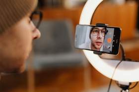 arts entrepreneurs: a photo of a young man wearing glasses and an orange beanie, recording himself on his phone in front of a professional lighting set-up.
