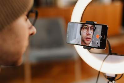 arts entrepreneurs: a photo of a young man wearing glasses and an orange beanie, recording himself on his phone in front of a professional lighting set-up.