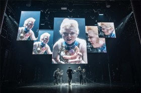 An image from the Broadway production of The Picture of Dorian Gray, with Tony-Award winning actor Sarah Snook peering into a camera and being broadcast across multiple screens.