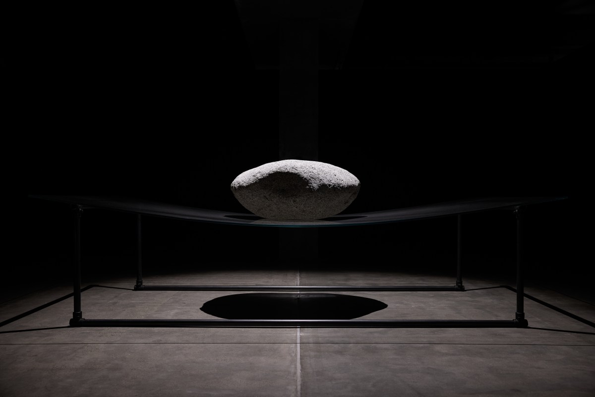 A large gray boulder sits atop a thin sheet of glass, bending it under teh pressure. Four stand legs are balancing the glass on each side, and the rest of the room is dark. 