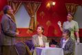 Set in a red carriage room with two women seated in a table, a woman and a man standing..