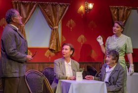 Set in a red carriage room with two women seated in a table, a woman and a man standing..