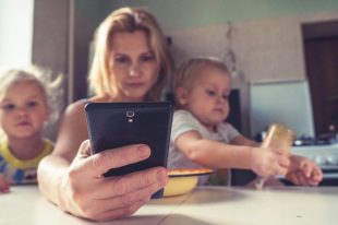 A photo of a mother managing two toddlers at a kitchen bench while also looking at her phone. Possibly a creative professional attempting to balance many side hustles.
