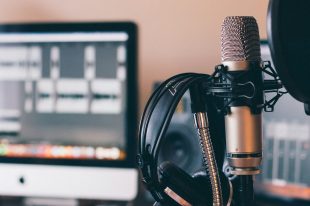 A set of headphones hangs over a microphone, in front of a computer where someone is editing a podcast.