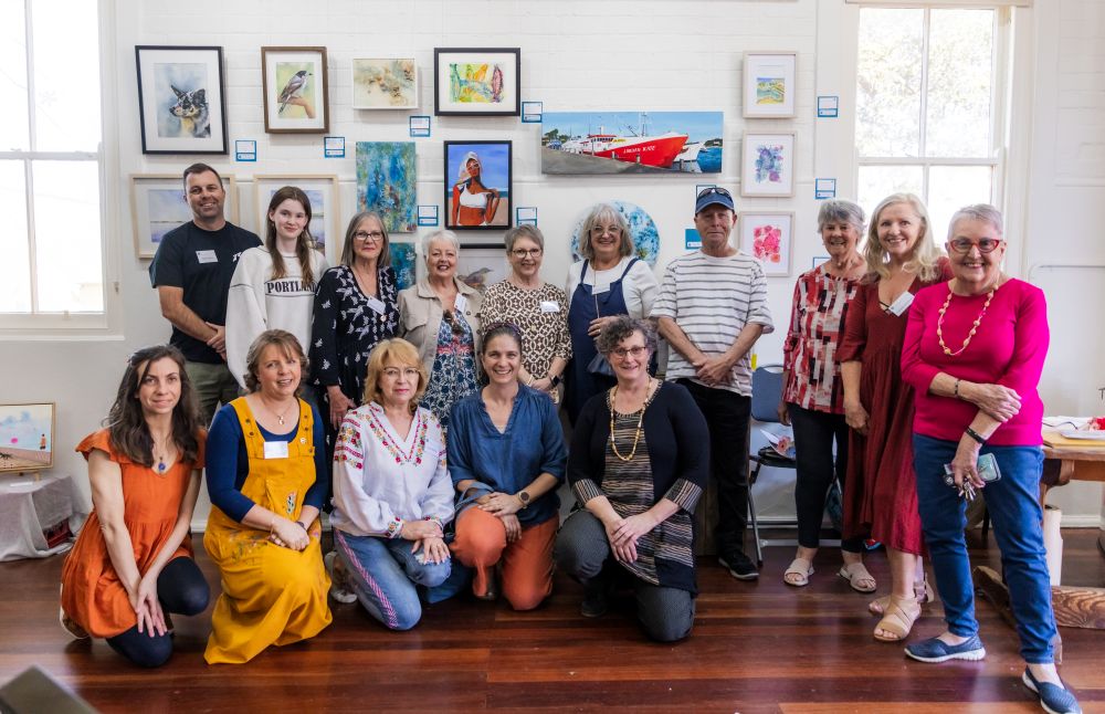 Fifteen artists, ranging in age, pose in front of a selction of artworks shown in Shellharbour Art Society's 2024 exhibition. Image supplied by Regional Arts Development Organisation South Coast Arts.