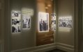 Black and white photographs of varying sizes arranged on a wall next to small text. Each of the photographs has a spotlight.