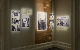 Black and white photographs of varying sizes arranged on a wall next to small text. Each of the photographs has a spotlight.