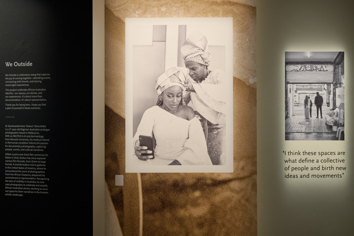 A black and white photo of an older African woman fixing a Nigerian head wrap on a younger woman while she is looking at herself through a phone. There is a small heading 'We Outside' to the left side of the wall. 