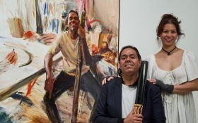 The artist Loribelle Spirovski (a young woman with brown hair in an up do smiling) with her hand on the shoulder of her sitter, William Barton (a middle-aged Aboriginal man holding a didgeridoo) in front of a large portrait painting of Barton sitting at in a room with a similar position.