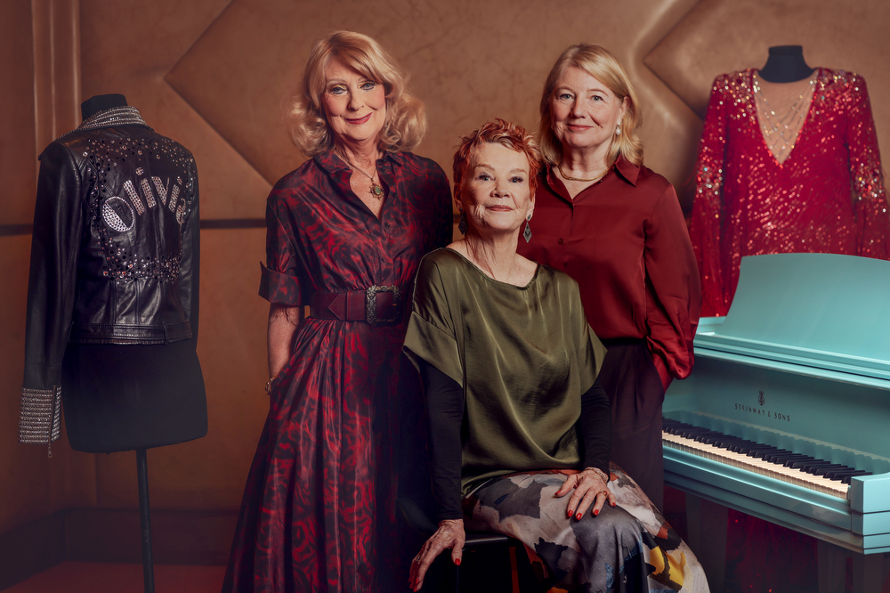 Three mature, fair-skinned female philanthropists pose together with objects recently acquired for the Australian Performing Arts Collection: a black leather jacket bedazzled with sequins, some of which spell out the word 'Olivia'; a glittering red gown, and a blue piano.