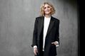 Ebony Bott, the incoming Artistic Director of Brisbane Festival. The photo shows a confident, fair-skinned woman with lightly curled, shoulder-length blonde hair smiling at the camera. She wears a charcoal-coloured jacket and pants over a white shirt.
