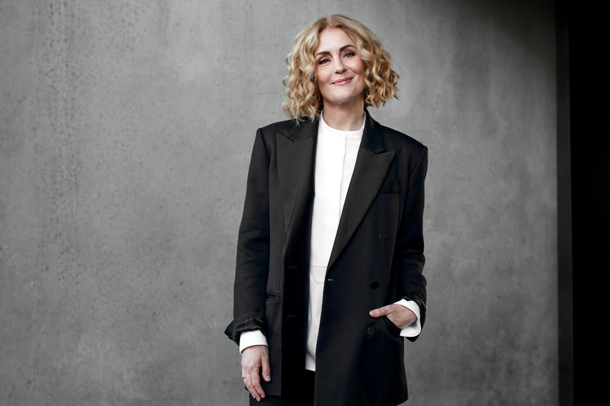 Ebony Bott, the incoming Artistic Director of Brisbane Festival. The photo shows a confident, fair-skinned woman with lightly curled, shoulder-length blonde hair smiling at the camera. She wears a charcoal-coloured jacket and pants over a white shirt.