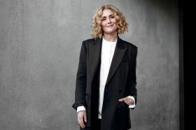 Ebony Bott, the incoming Artistic Director of Brisbane Festival. The photo shows a confident, fair-skinned woman with lightly curled, shoulder-length blonde hair smiling at the camera. She wears a charcoal-coloured jacket and pants over a white shirt.