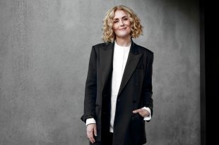 Ebony Bott, the incoming Artistic Director of Brisbane Festival. The photo shows a confident, fair-skinned woman with lightly curled, shoulder-length blonde hair smiling at the camera. She wears a charcoal-coloured jacket and pants over a white shirt.