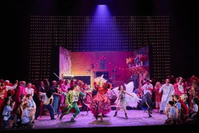 Colourful cast scene, with a crowd looking on at two women and a man dancing in the middle. Carmen.