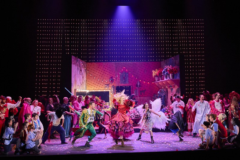 Colourful cast scene, with a crowd looking on at two women and a man dancing in the middle. Carmen.