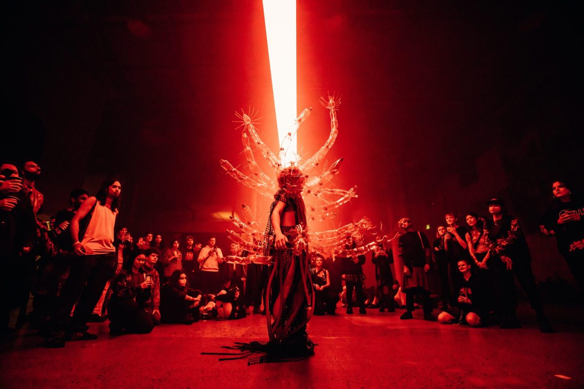 An artist dress in a costume with cactus-like limbs sticking out of their back surrounded by a circle of onlookers. The room is awash in red light. SOFT CENTRE 2022.