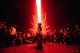 An artist dress in a costume with cactus-like limbs sticking out of their back surrounded by a circle of onlookers. The room is awash in red light. SOFT CENTRE 2022.