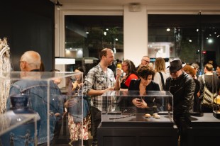 Defunding success in the arts. ADC MAKE Award Special Event. A group of people mingle around some glass/perspex boxes containing art.